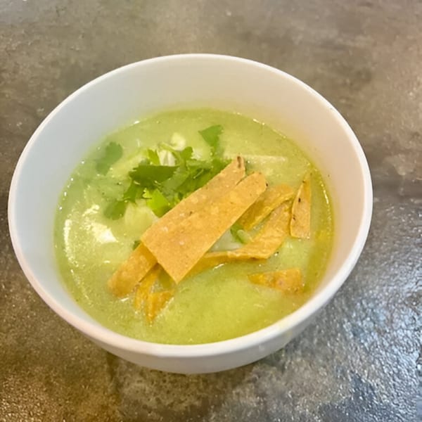 A white bowl of Falafel Taco's green soup, garnished with cilantro and tortilla strips.