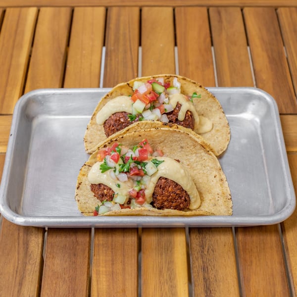 Two Falafel Tacos on a metal tray, topped with pico de gallo and a creamy sauce.