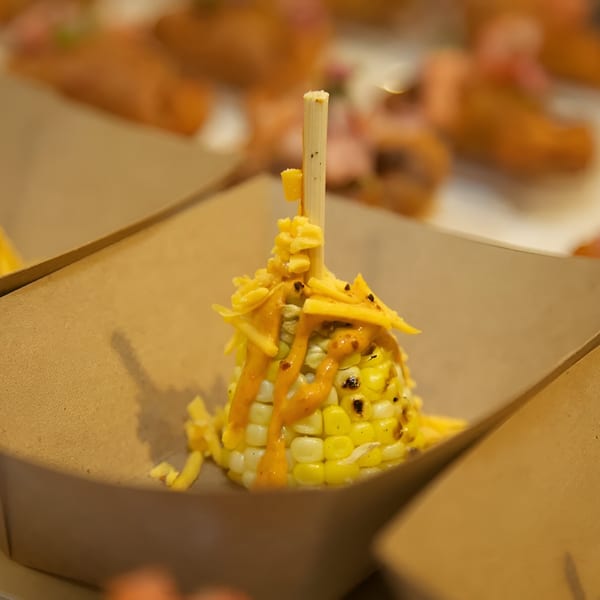 Grilled corn on the cob with melted cheese and sauce, served in a brown paper tray.